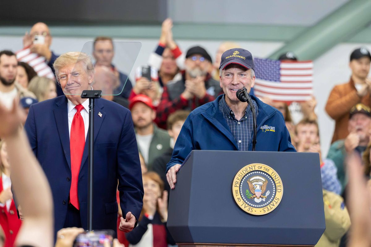 Senator Dan Sullivan speaks at a microphone in Wasilla while President Trump, in a navy suit and red tie, stands beside him. Both faces are in the frame.
