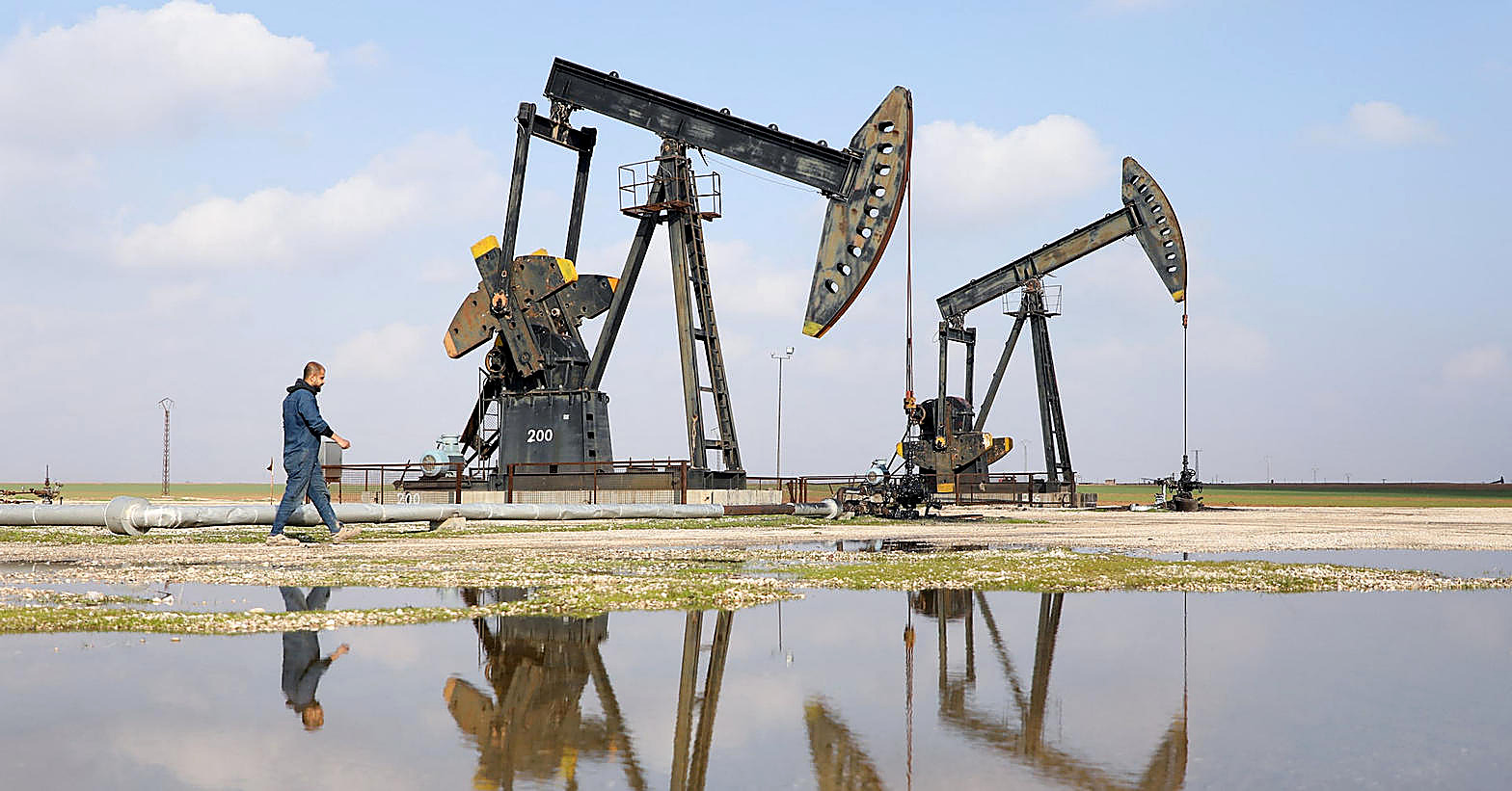 A man in blue walks past two pumpjacks in a field in Syria, their reflections in a pool of water.