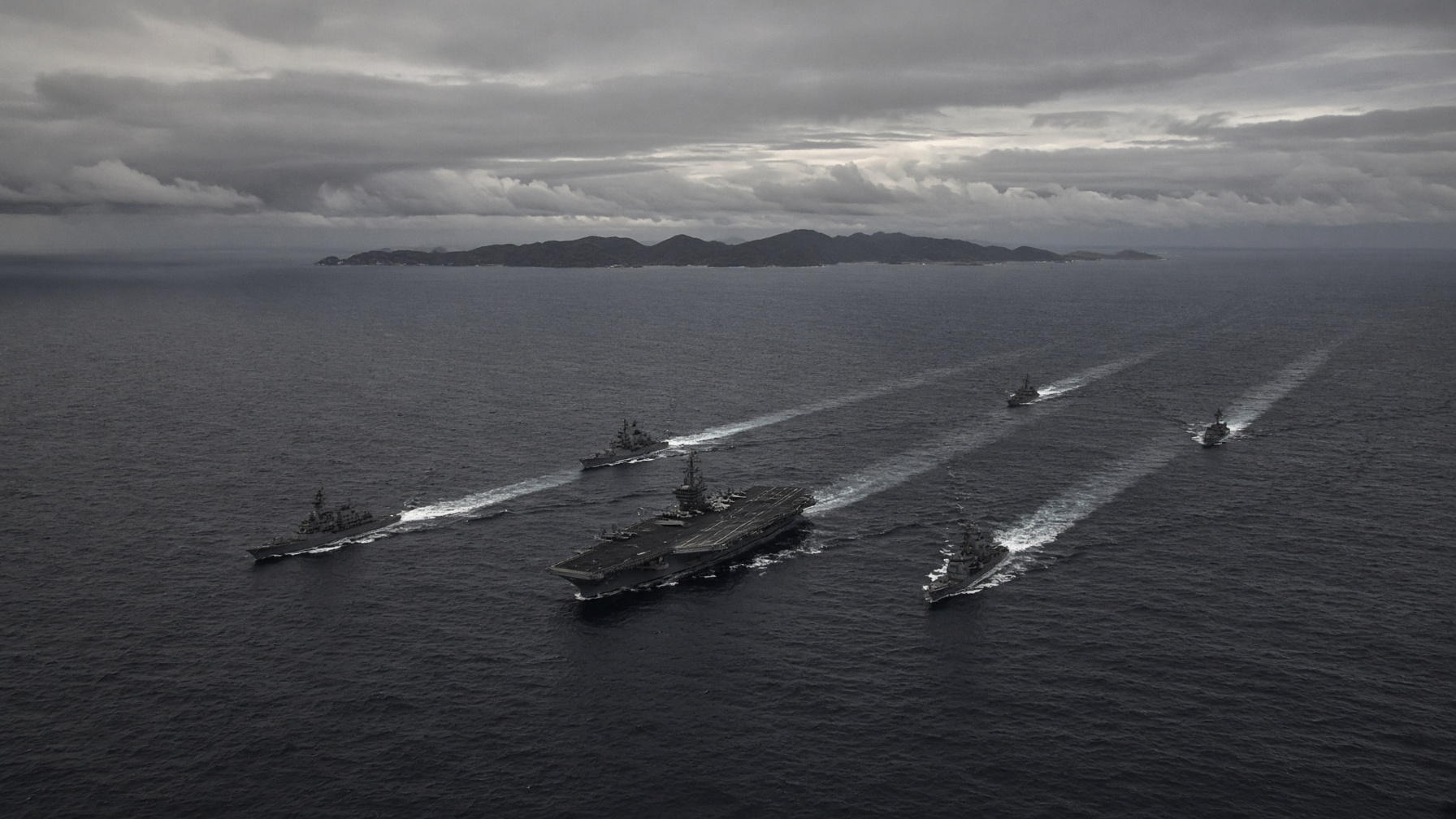 An aircraft carrier strike group leaving long white wakes across a gray Pacific under overcast sky.