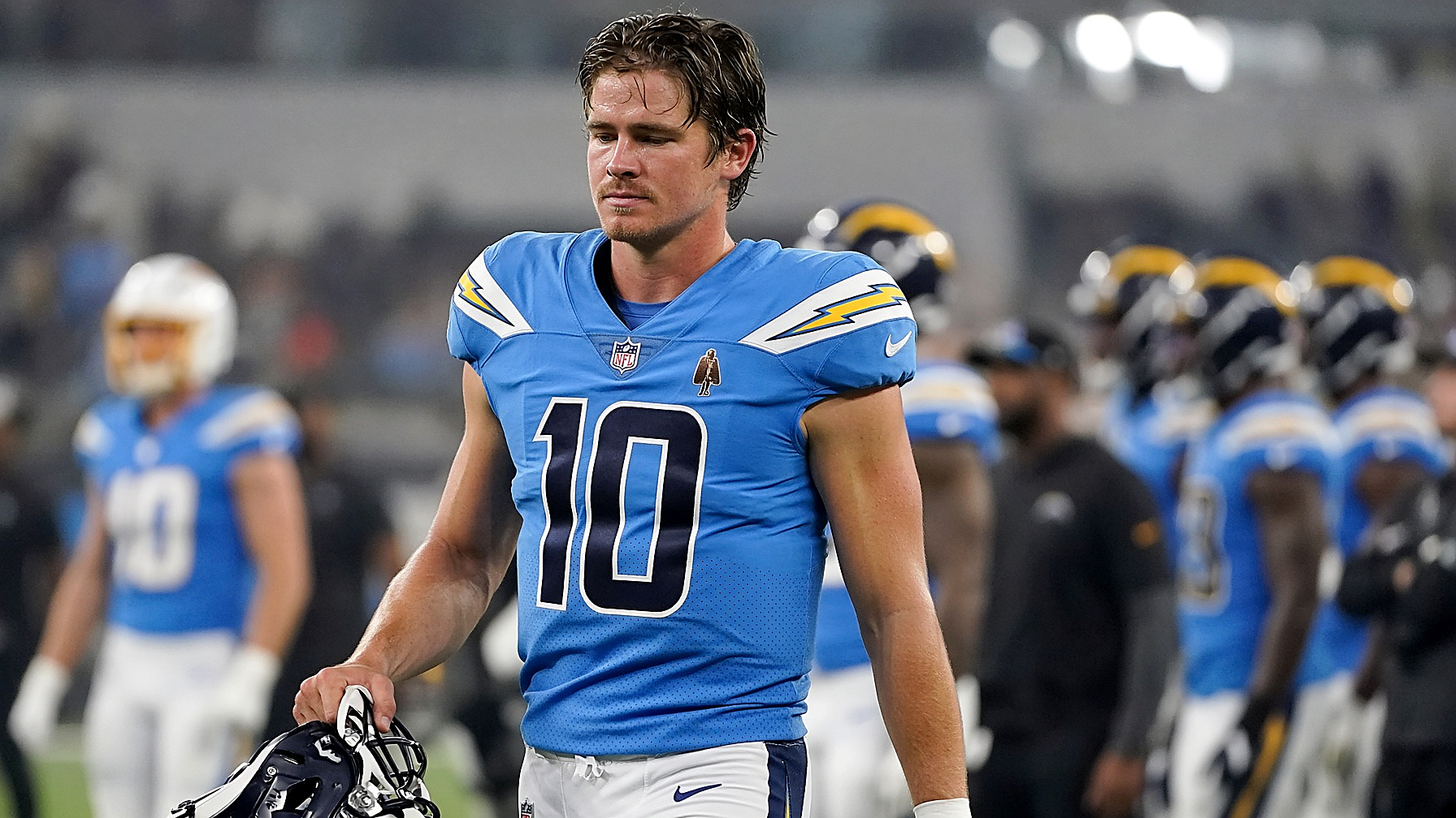 A Chargers quarterback in powder blue on the sideline, helmet in hand, stadium lights, in color. Person plate.