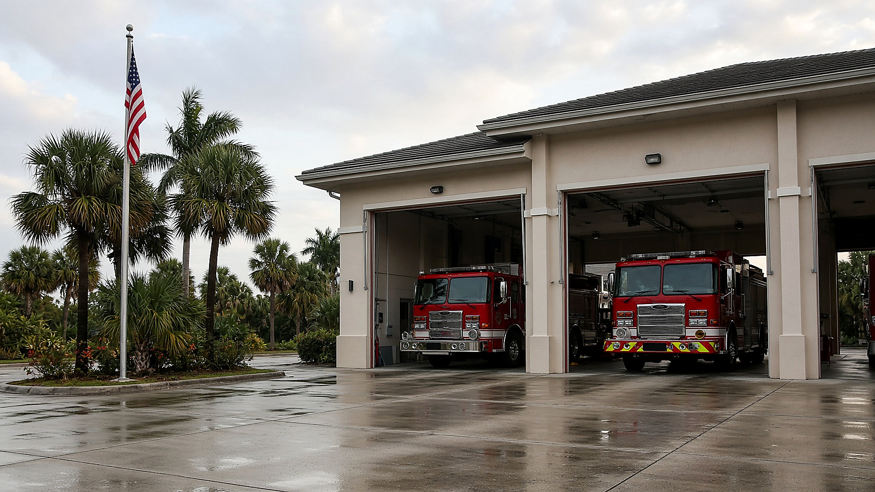 A Florida fire station at morning, two red engines in open bays, palms and flag, in color. No figures in the frame.