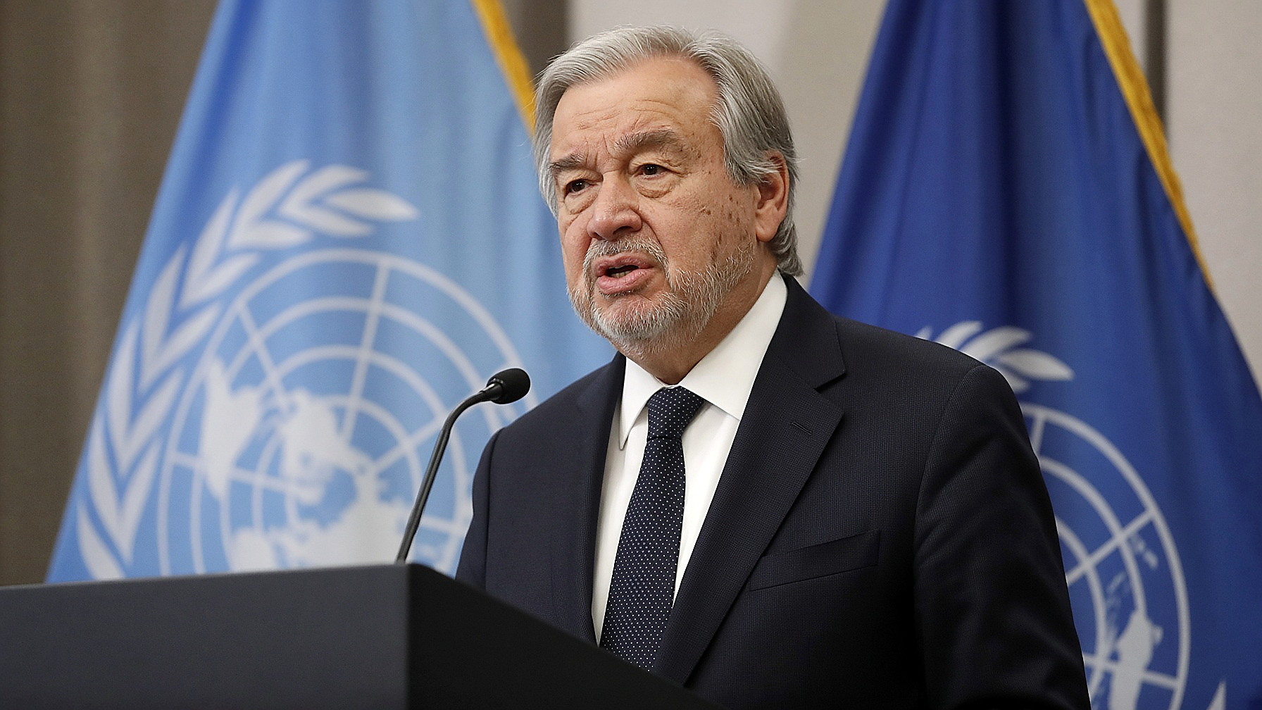 U.N. Secretary-General António Guterres at the United Nations podium, flags behind him, in color. Person plate.