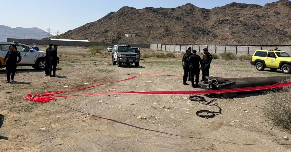 Officials stand over missile debris marked by a red ribbon on open ground near Mecca, with mountains behind them.