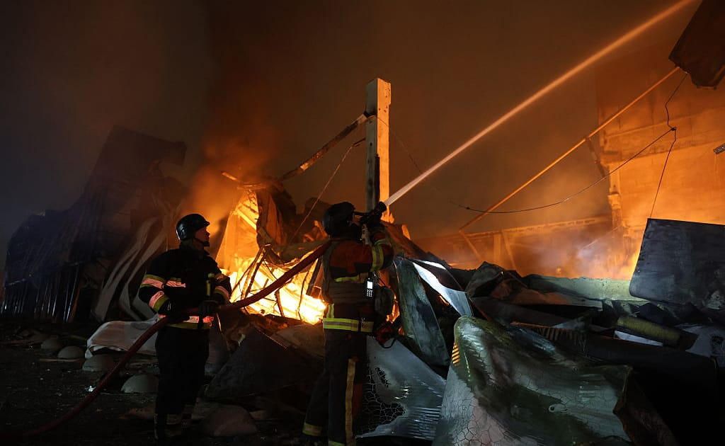 Two firefighters in helmets pour a hose onto a fire in a collapsed building at night. A third firefighter stands in the smoke.