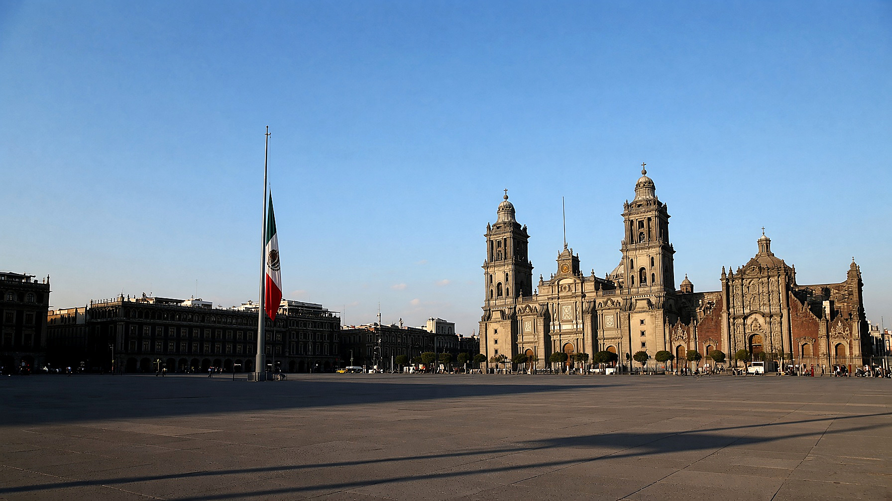 The Mexican flag at half-mast over an empty Zócalo, cathedral behind, Saturday morning, in color. No figures in the frame.