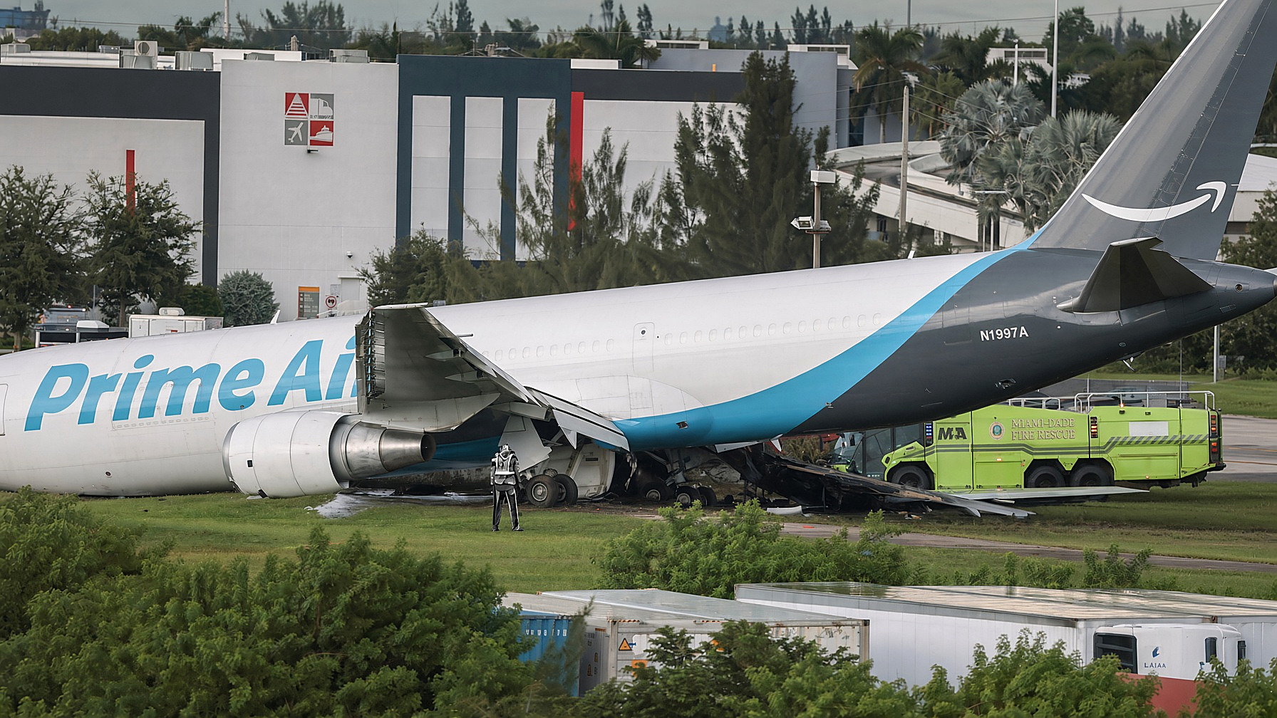 Prime Air Boeing 767 N1997A on the grass at Miami International, torn fuselage, lime Miami-Dade fire truck in color; figures at the nose in black and white. Scene photograph, Sunday, Sept. 6, 2026.