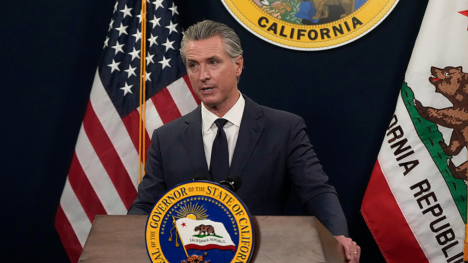 Gov. Gavin Newsom at a California podium, full color, navy suit, state seal. Wire photograph.