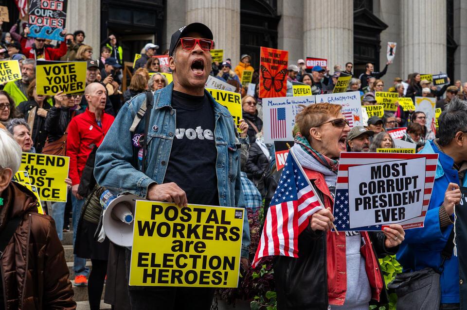 A man shouts into the crowd at a No Kings rally, a megaphone in one hand and a sign calling postal workers election heroes.