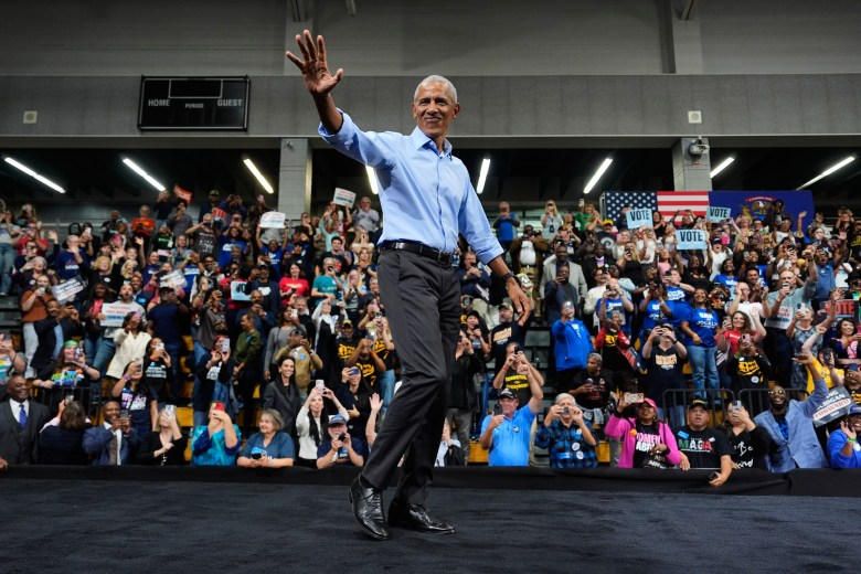 Barack Obama waves in a light blue shirt on a dark stage, a crowd with signs behind him at a high school gym in Detroit.