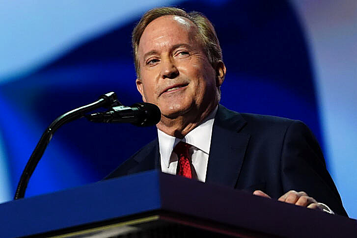 Ken Paxton at a podium, full color: navy suit, red tie, blue backdrop. Wire photograph.