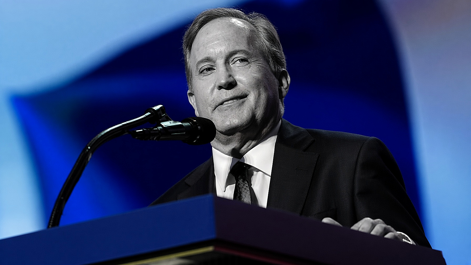 Ken Paxton at a podium: the figure in black and white — suit, tie, hands — the blue backdrop and lectern in color. Wire photograph.