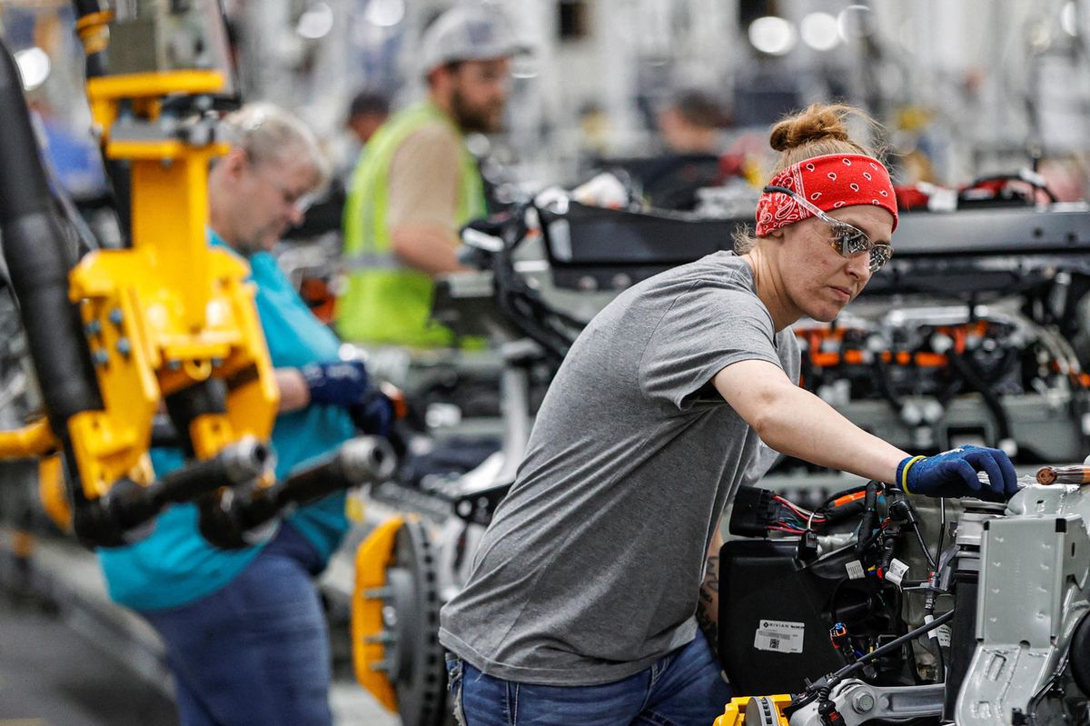 A worker in a red bandana and safety glasses reaches into a vehicle frame on a Rivian assembly line.