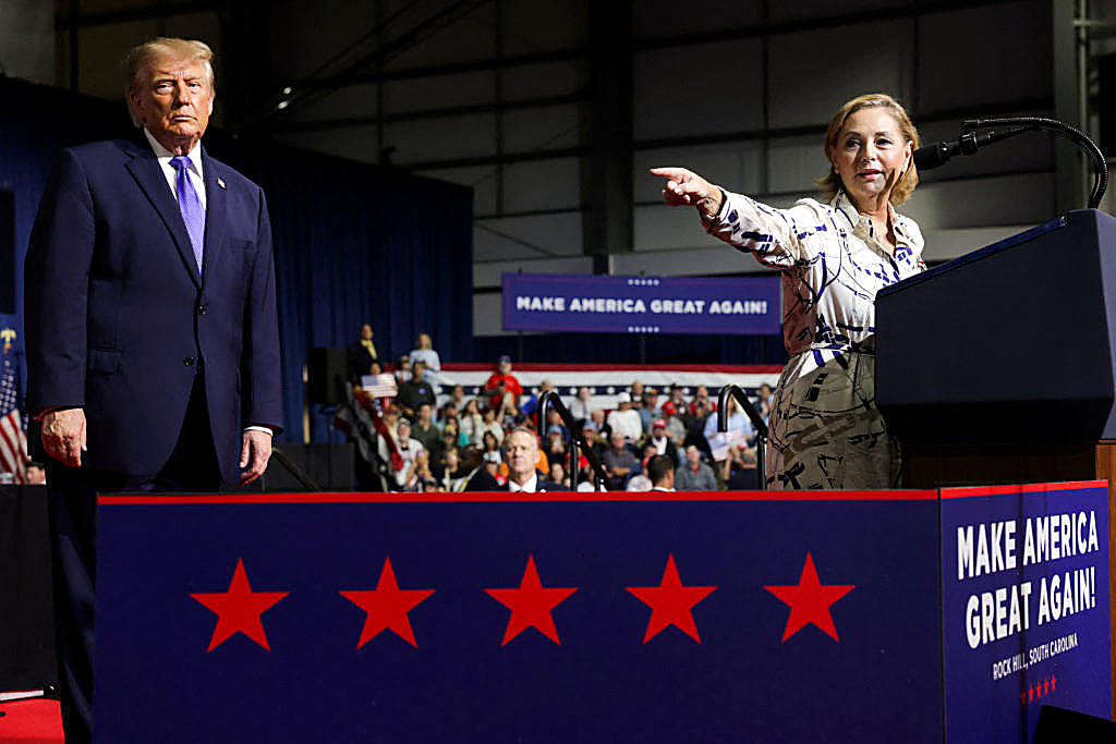 Sen. Darline Graham points from the lectern while President Trump stands beside her. Thursday, Oct. 29, Rock Hill Sports and Event Center. Reuters. Both faces are in the frame. The lectern reads Rock Hill, South Carolina. It is not Florida.