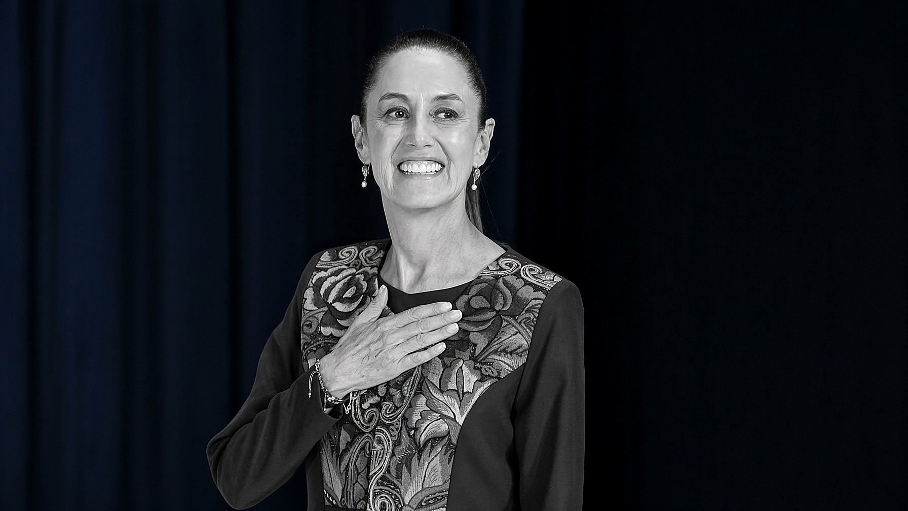 President Claudia Sheinbaum, hand on her chest: the figure in black and white; the curtain behind her in color. Wire photograph.