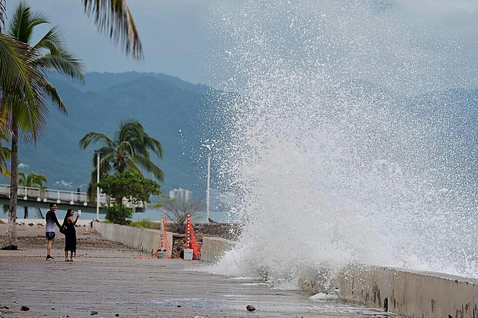 A wave from Hurricane Simon breaks over a Pacific seawall as two people watch from the walk.