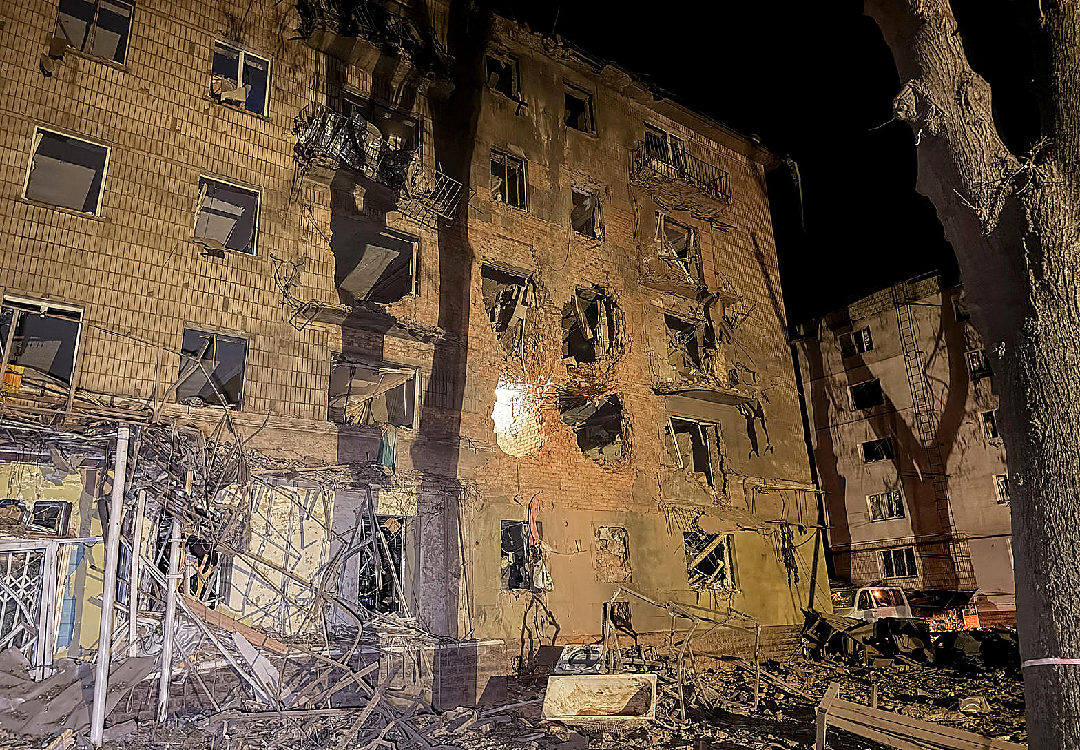 A Ukrainian apartment block at night, windows blown, rubble in the yard, in color. No figures in the frame.