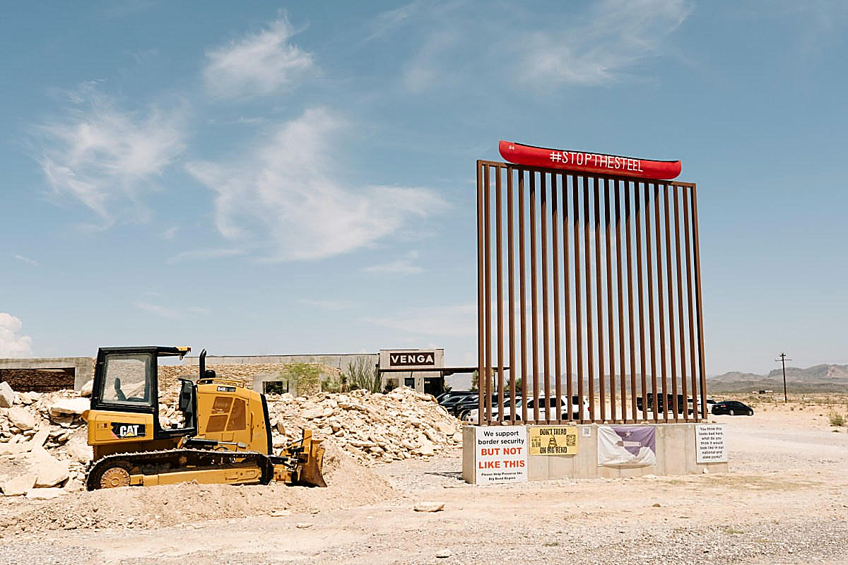 A yellow bulldozer and a sample wall panel in the Big Bend desert, a canoe reading STOP THE STEEL, in color. No figures in the frame.