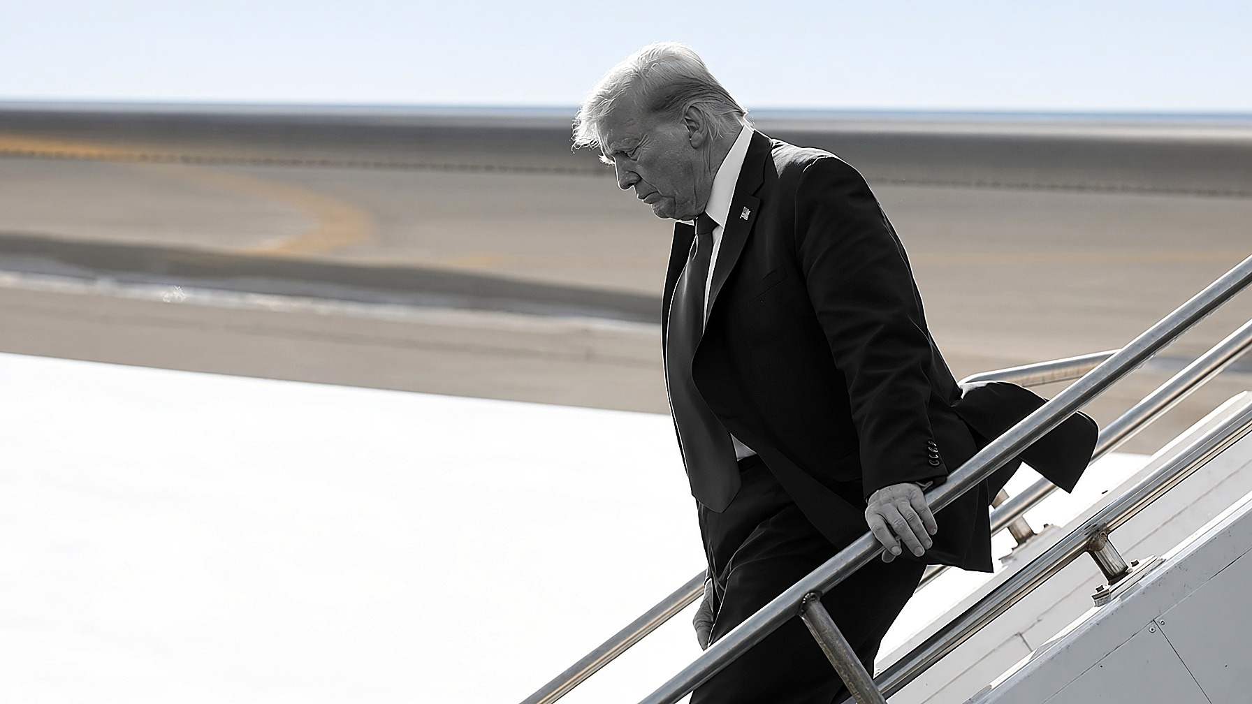 President Donald Trump descending Air Force One stairs, looking down, right hand on the rail. Figure in black and white; tarmac, sky and metal stairs in color.