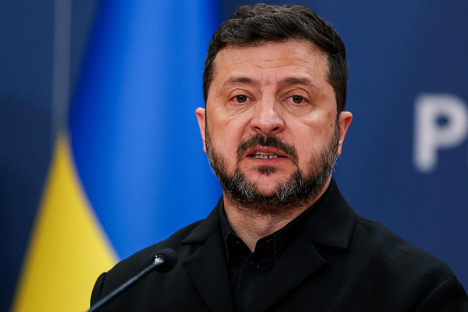 President Volodymyr Zelenskyy at a microphone, Ukrainian flags behind him, in color. Person plate.